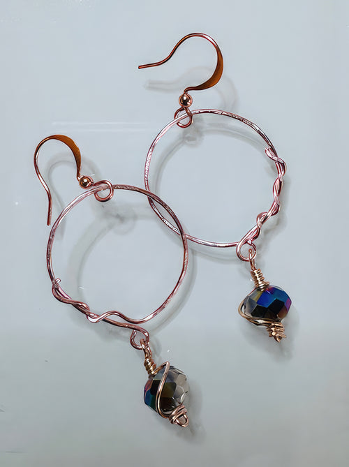 A pair of handcrafted earrings featuring wrapped copper wire and Czech glass beads in shades of purple, green, and clear, with a copper hoop design.
