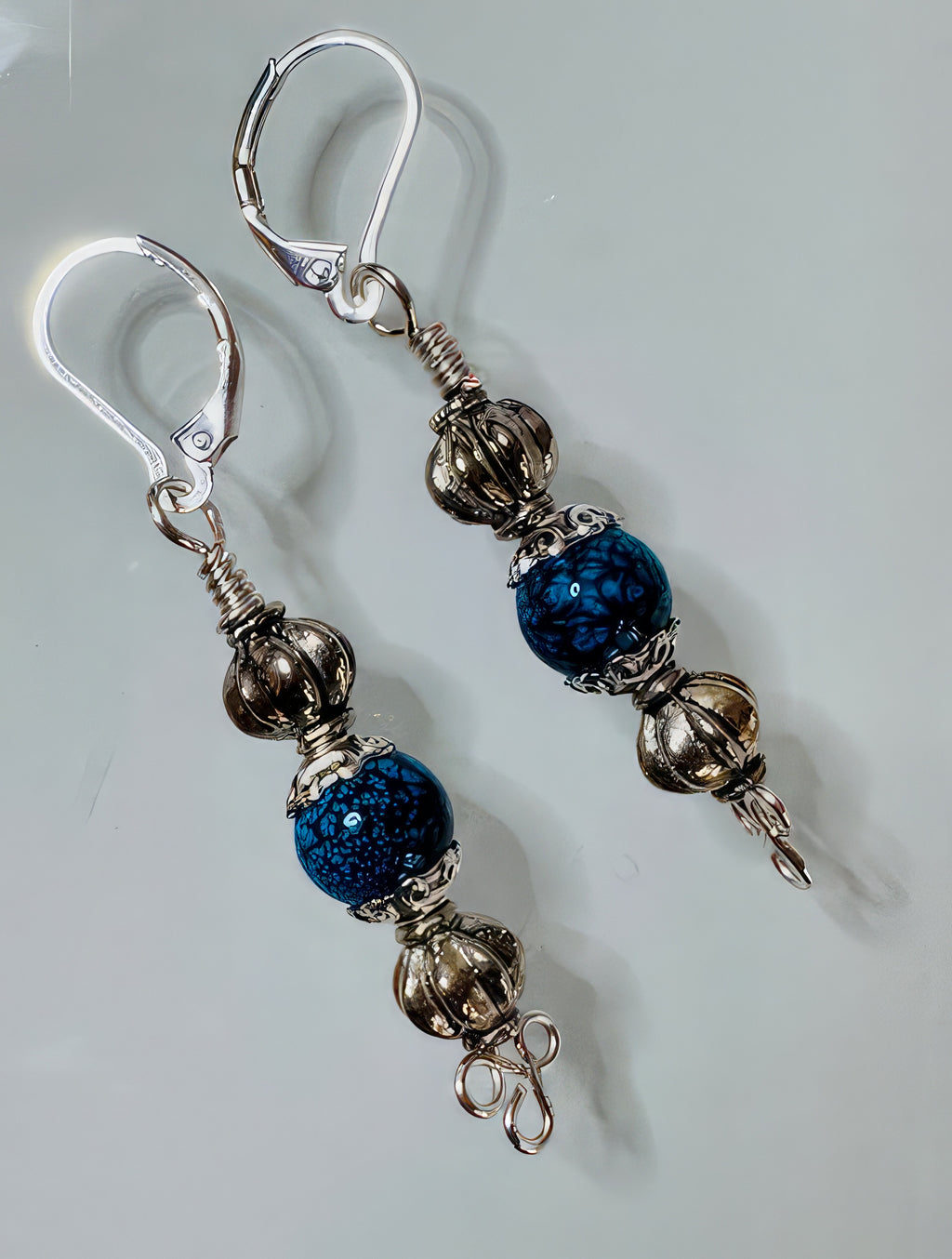 A pair of earrings featuring a handmade clover wire base with highly textured antiqued silver-tone beads, bead caps, and a royal blue stone, finished with hypoallergenic leverback ear wires.