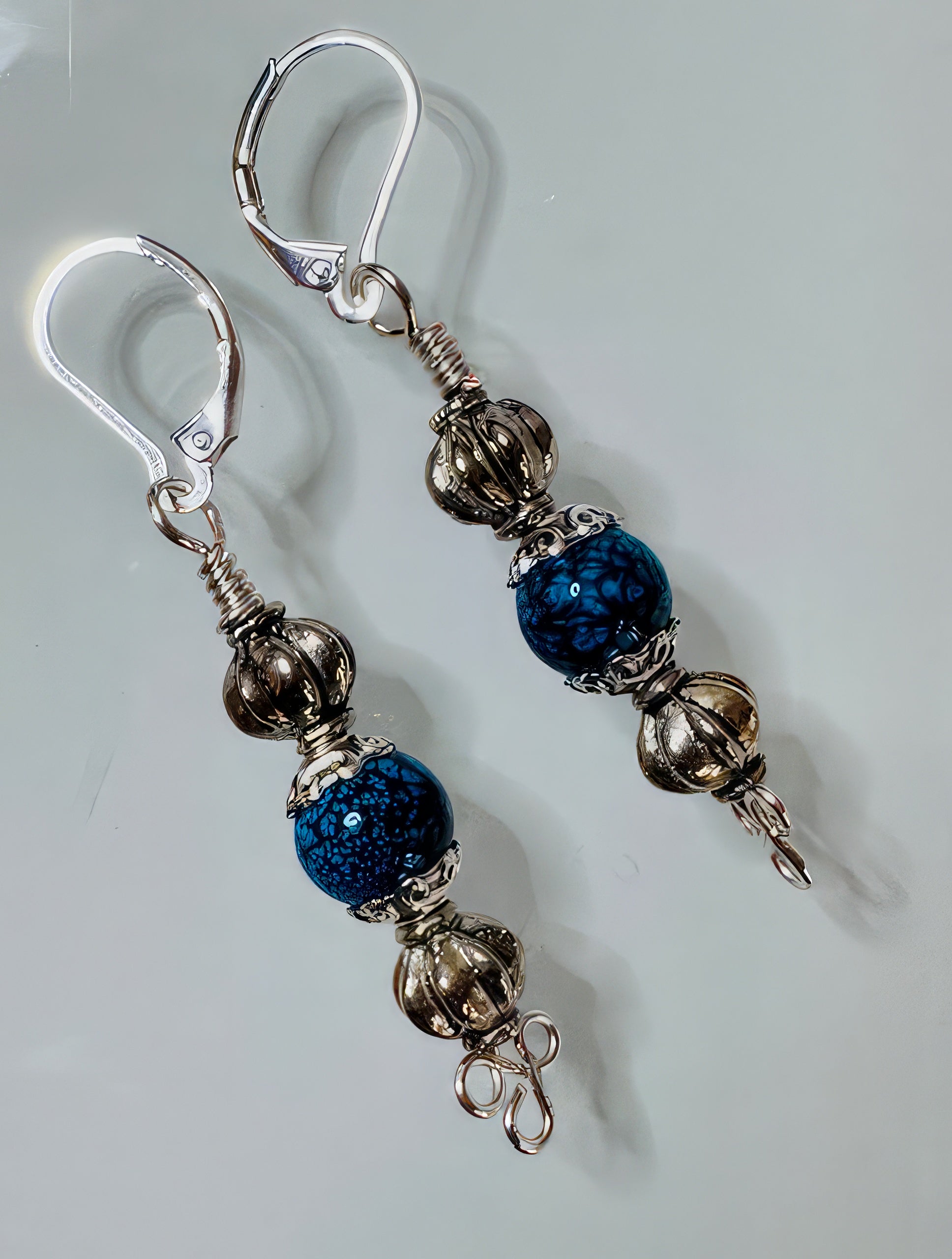 A pair of earrings featuring a handmade clover wire base with highly textured antiqued silver-tone beads, bead caps, and a royal blue stone, finished with hypoallergenic leverback ear wires.