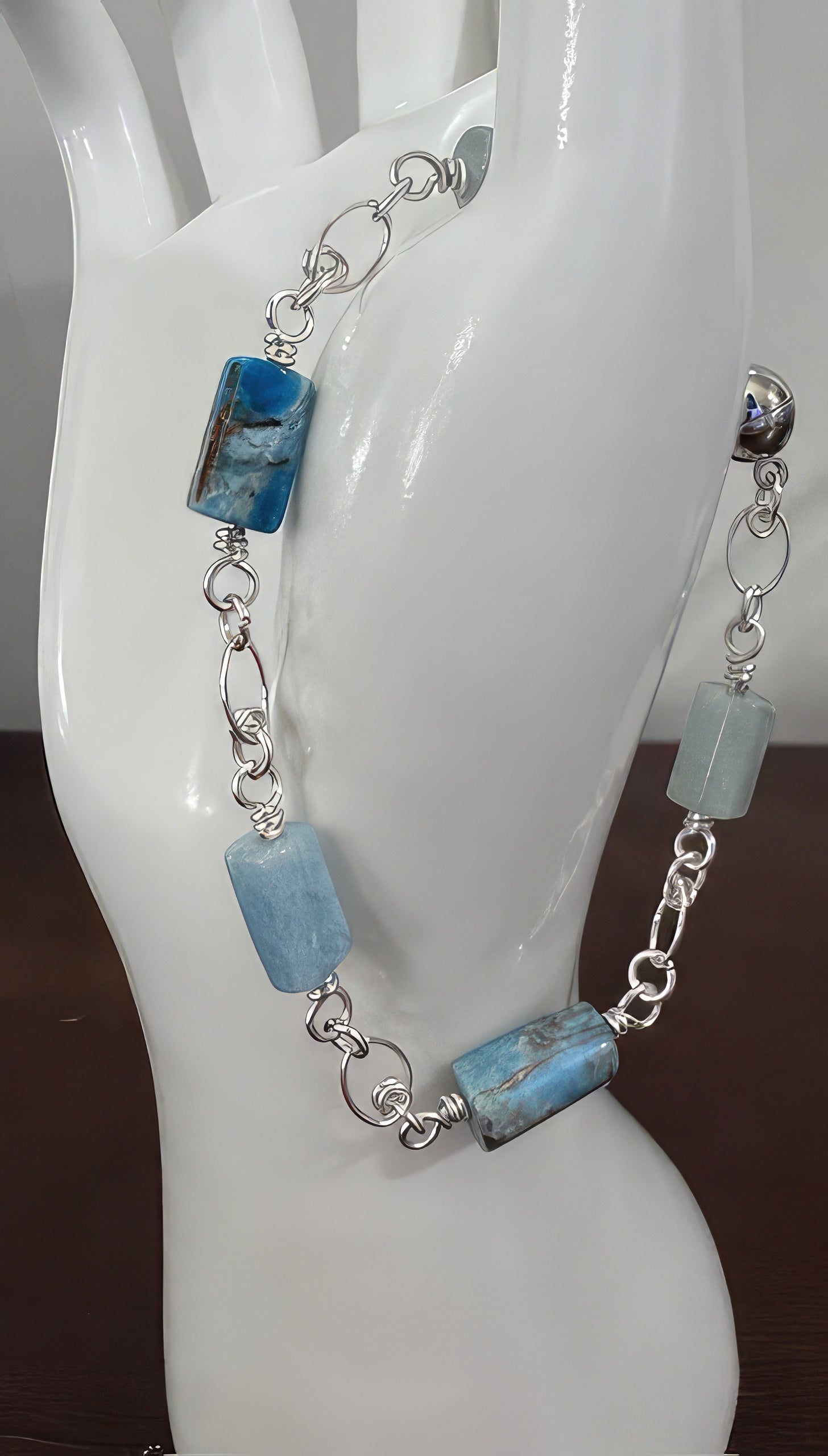 A bracelet with blue tumbled stones and silver-colored oval links, displayed on a white mannequin hand.