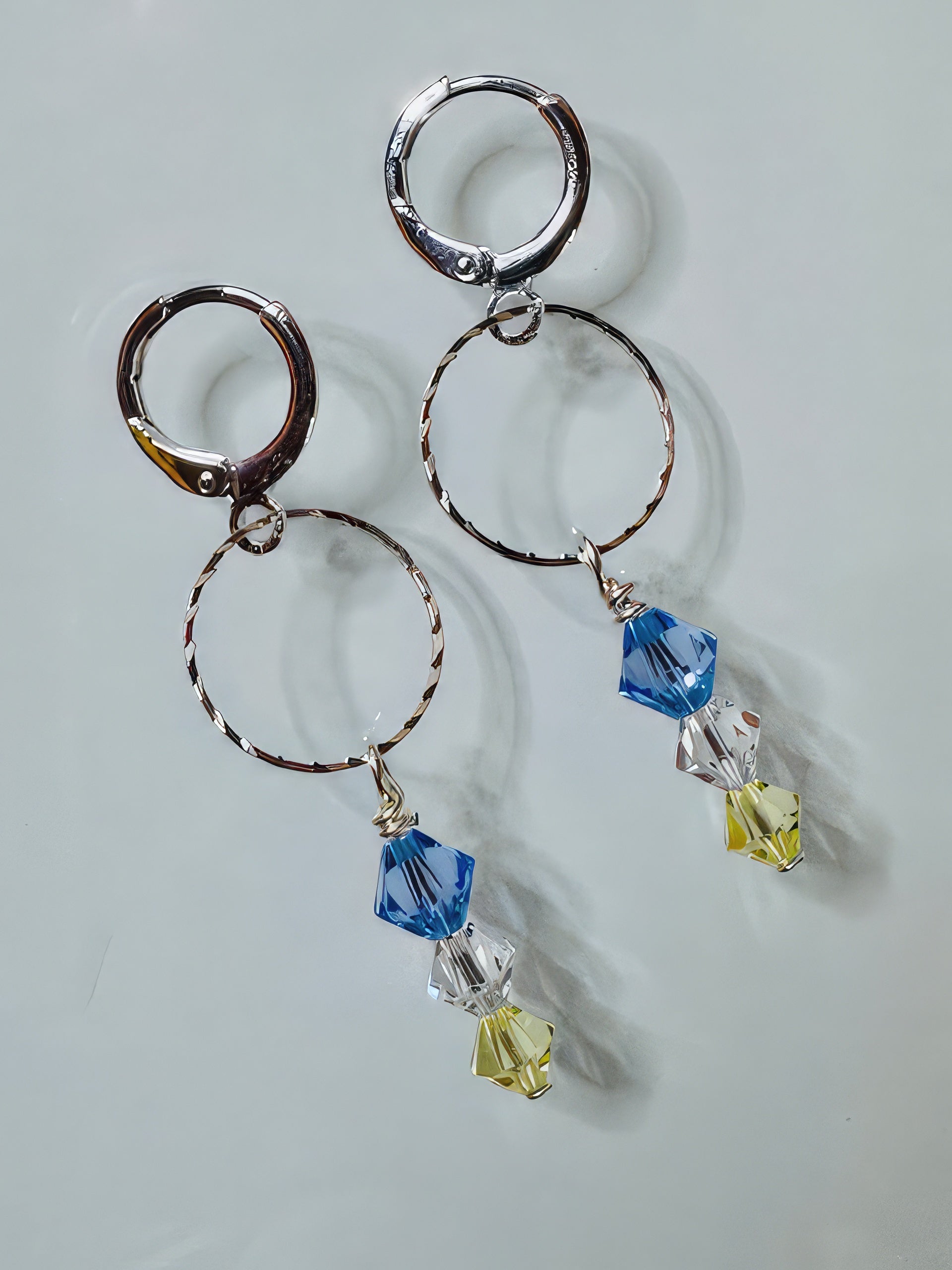 A pair of earrings featuring silver-filled faceted hoops and suspended pastel Swarovski crystals in blue, clear, and yellow colors with petite leverback earwires.