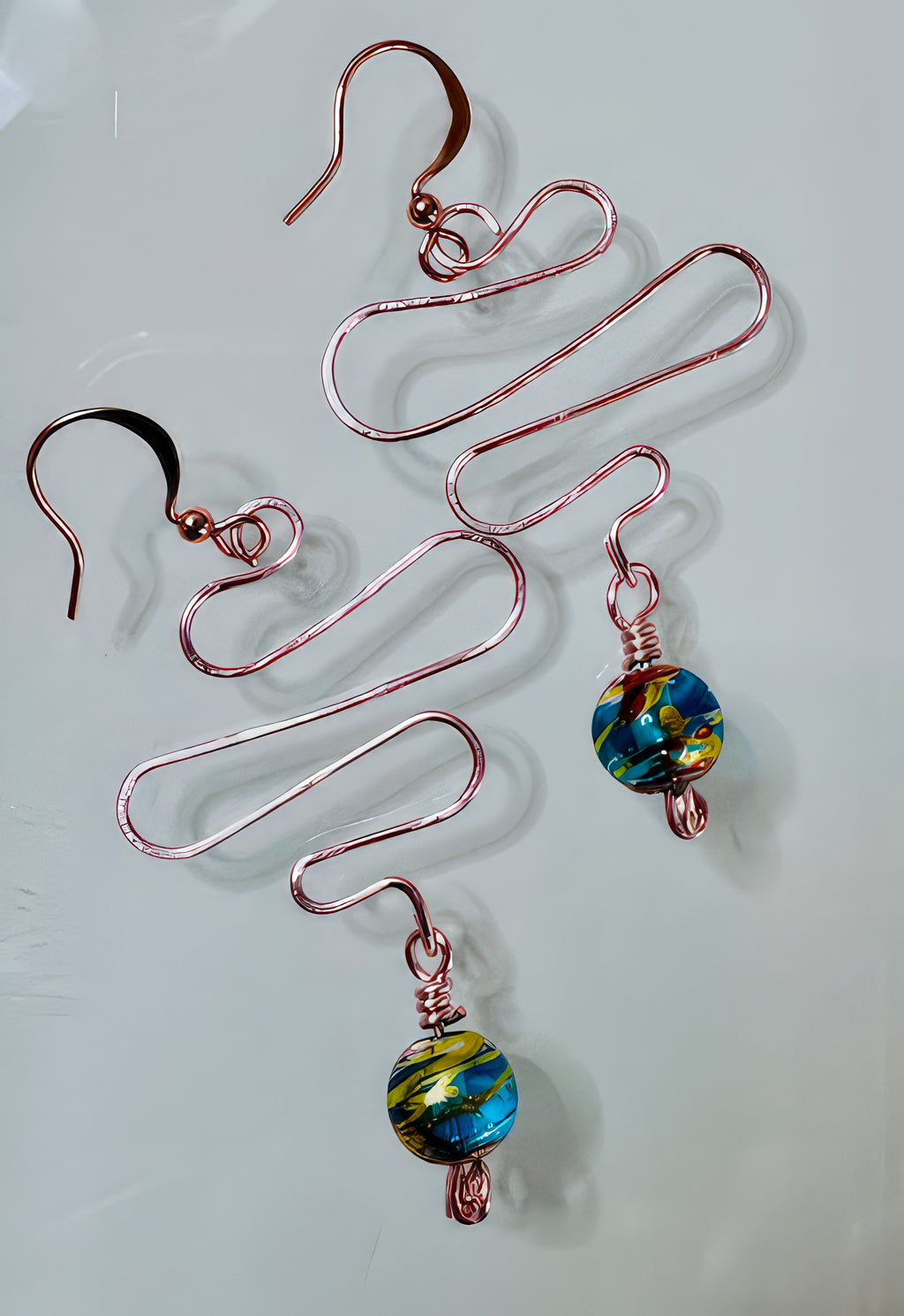 A pair of earrings with hand formed copper wire and a teal blue with hints of gold painted glass bead, suspended from copper core ear wires with a non-tarnish coating.