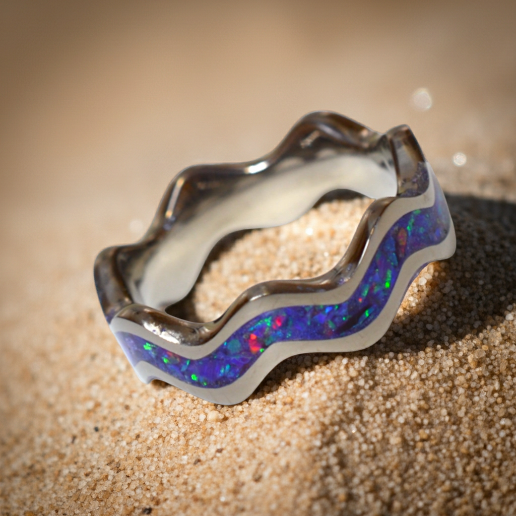 Handmade wave-shaped titanium ring with a handcrafted crushed opal inlay, photographed on sand to highlight its ocean-inspired design.